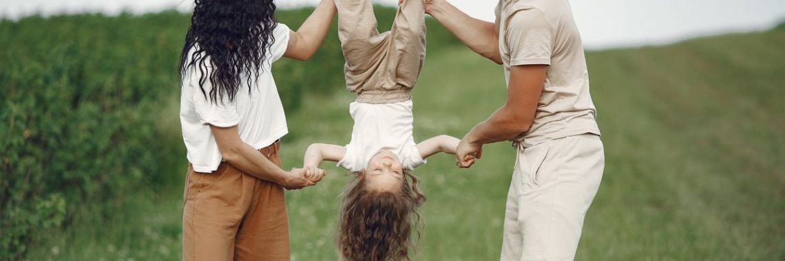 A joyful family enjoys outdoor play, lifting their child upside down in a green field.