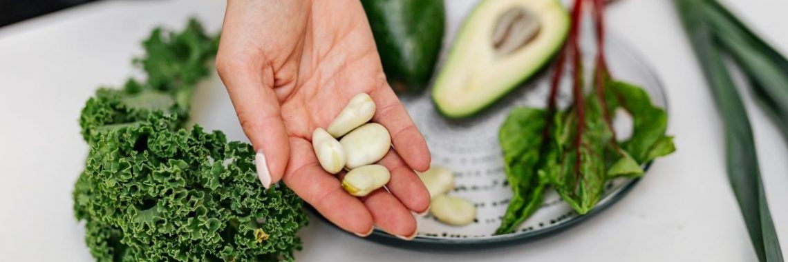Close-up of a hand holding garlic cloves over a plate with fresh vegetables, including kale and avocado.